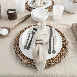 Minimalist table setting featuring linen napkins, a cotton tablecloth, and round rattan placemats on a neutral-toned dining table.