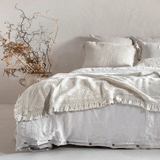 Minimalist natural bedroom interior featuring white linen bedding, textured cushions, fringed throw and large ceramic vase.