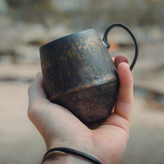 Handmade stoneware mug with a bronze metallic glaze, inspired by wabi-sabi aesthetics, displayed in holding hand.