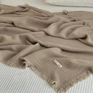 Nori cotton muslin throw in taupe with soft fringed edges draped over a bed