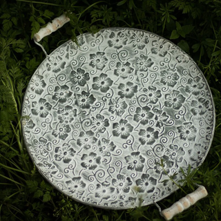 Round floral embossed galvanized iron tray, a stylish metal serving plate with handles perfect for kitchen tableware. This durable serving plate features intricate embossing, adding a touch of elegance to your dining experience. Ideal for both practical use and natural home decor, it enhances your table setting with rustic charm and versatility.