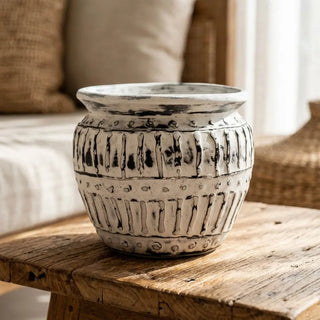 A round ceramic planter pot with a textured, hand-painted off-white and black rustic pattern, sitting on a wooden table in a cozy living room setting.
