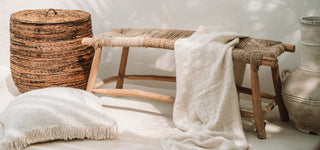 Cotton and linen blankets and cushions, rattan basket, rustic bench and large earthy ceramic vase styled in a neutral decor.