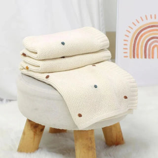 Bibi Cotton Knit Baby Blanket in soft white, folded neatly on top of rustic wooden stool.