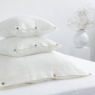 Stack of folded white linen pillows and duvet cover with coconut buttons, styled in a minimalist, scandi bedroom.