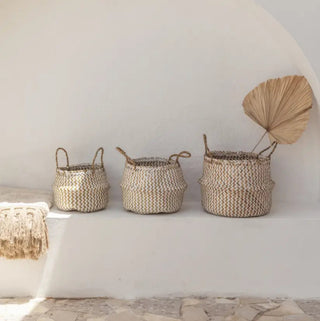Three handwoven seagrass baskets in natural and white on a plaster shelf with dried palm leaves