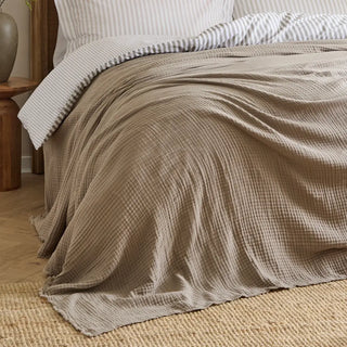 Nori latte cotton muslin throw spread over a bed with striped linen bedding